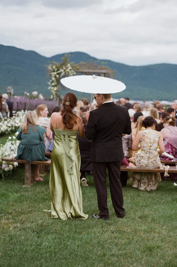 A couple standing holding a parasol at the back of the ceremony space on The Knoll. 