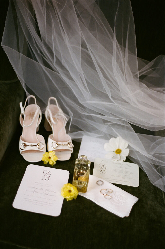 A flat lay of the couples invitations, the brides perfume, shoes, and veil, on a green velvet chair. 