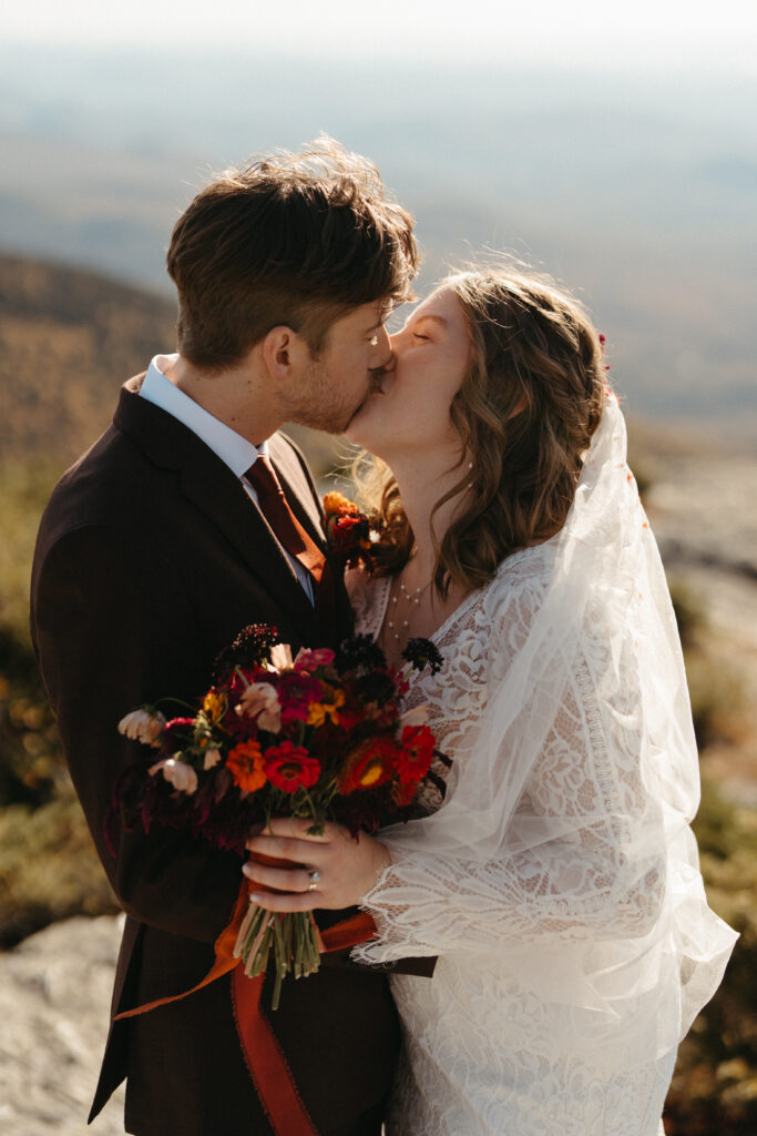 A couple kissing on top of Mount Mansfield holding a bouquet in their wedding attire. 