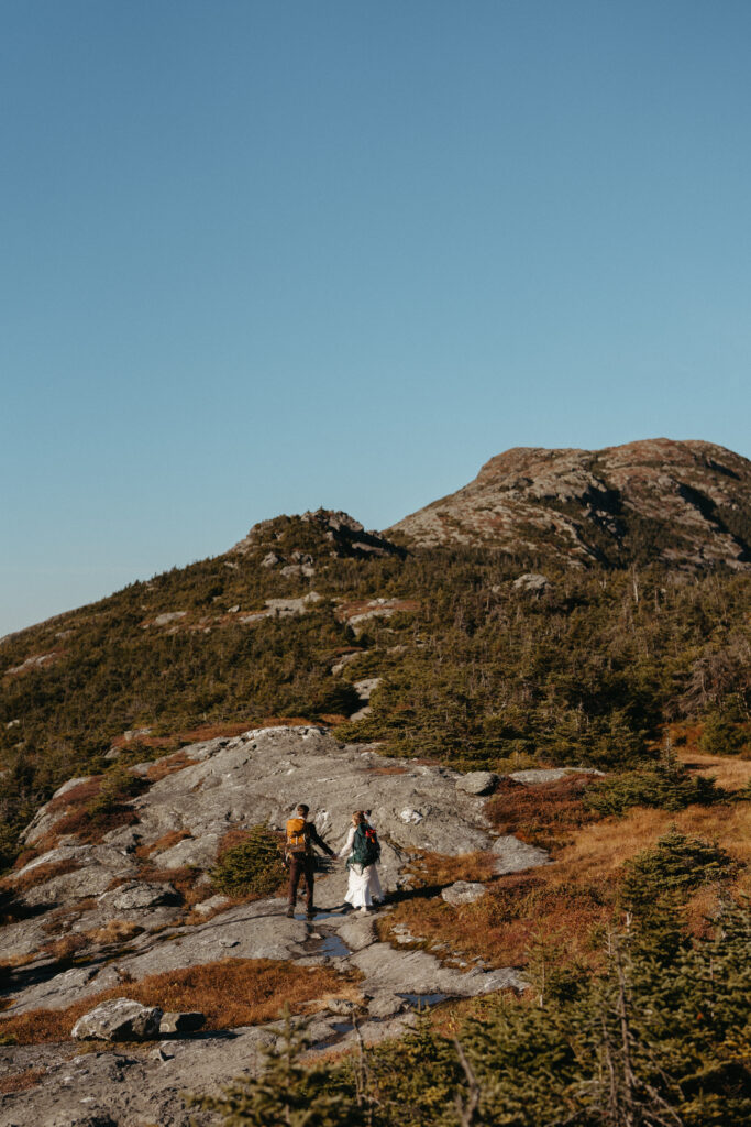The wedding couple walking up Mount Mansfield's granite slabs with their backpacks on. The couple is very far away from the camera and looks tiny against the vast landscape. 