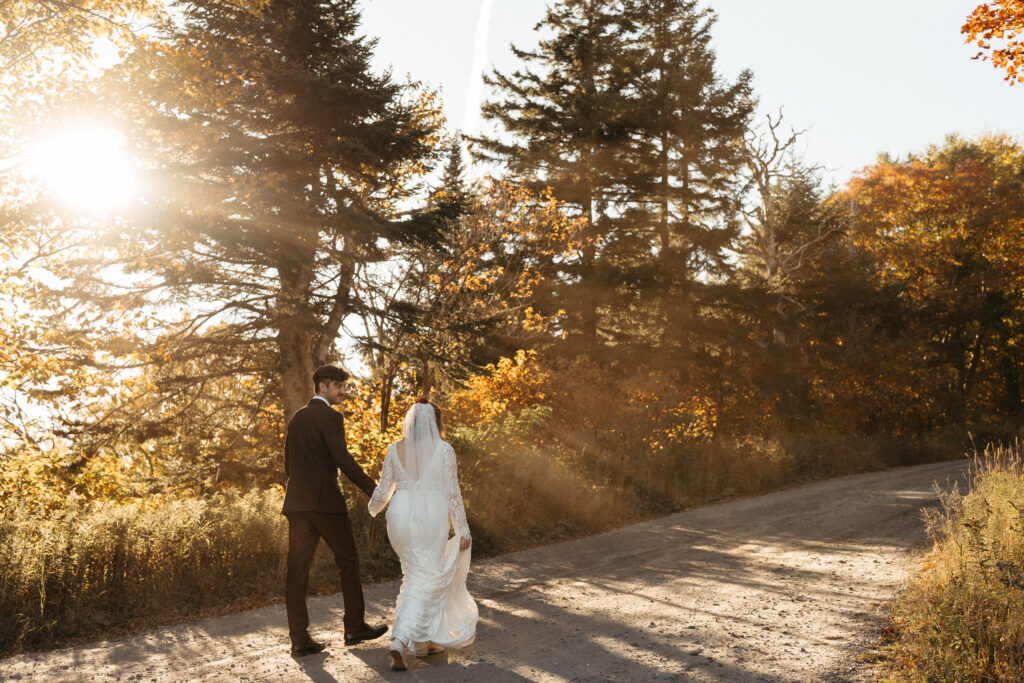 The light filtering through the trees and dust after a car drove down the Mount Mansfield Auto Toll Road. The couple is walking in the middle and the groom is looking at the bride. 